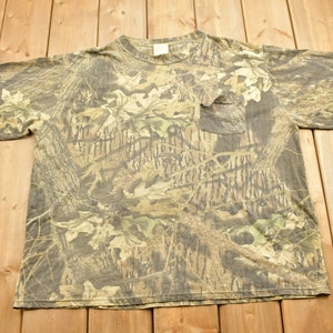 May include: A camouflage-patterned t-shirt with a pocket on the chest. The shirt features a blend of brown, green, and tan hues, mimicking a forest environment. The short-sleeved shirt is made of a soft material.