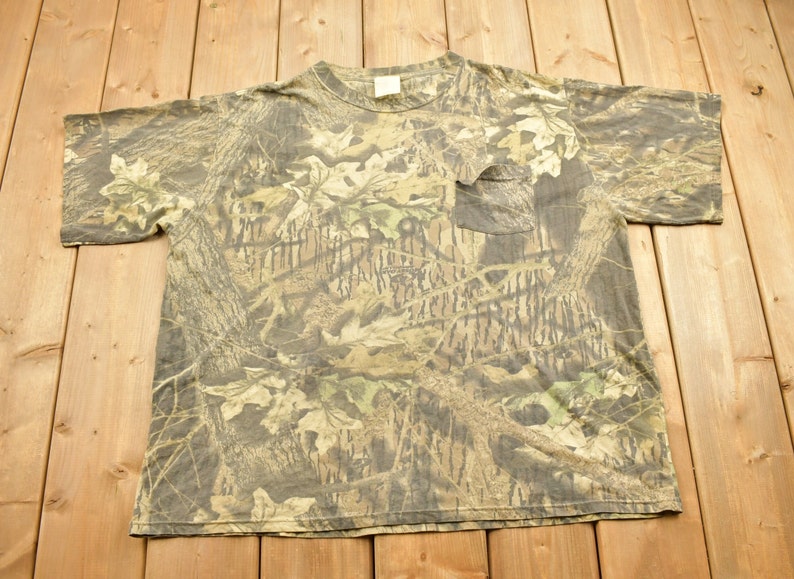 May include: A camouflage-patterned t-shirt with a pocket on the chest. The shirt features a blend of brown, green, and tan hues, mimicking a forest environment. The short-sleeved shirt is made of a soft material.