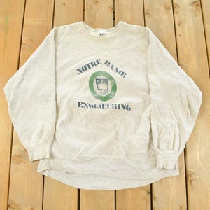 May include: A light gray sweatshirt with the words "NOTRE DAME ENGINEERING" printed on the front. The text is in a dark blue color, with a green and white emblem in the center. The sweatshirt is made of a soft material.