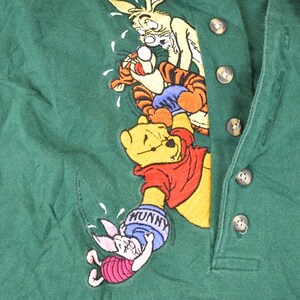 May include: Green collared shirt featuring an embroidered design of Winnie the Pooh and friends. The design includes Pooh Bear, Tigger, Rabbit, and Piglet. The shirt has a button-up placket and a tag.