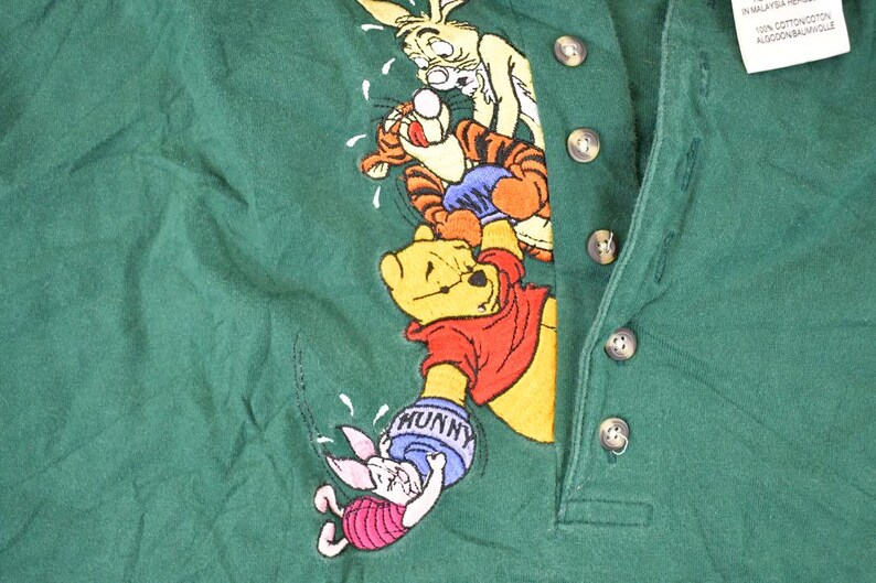 May include: Green collared shirt featuring an embroidered design of Winnie the Pooh and friends. The design includes Pooh Bear, Tigger, Rabbit, and Piglet. The shirt has a button-up placket and a tag.