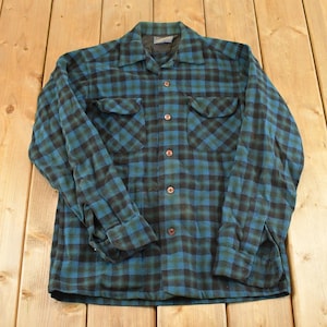 Puede incluir: Una camisa de cuadros vintage de manga larga en verde azulado y negro. La camisa tiene dos bolsillos en el pecho y una botonadura frontal. La tela tiene un aspecto suave y desgastado. La camisa está tendida sobre una superficie de madera.
