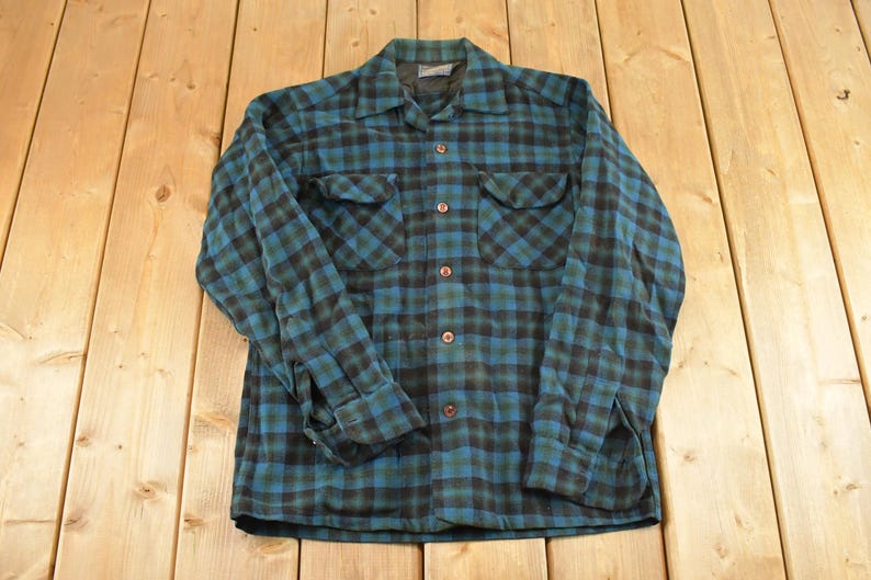 Op de afbeelding: Een vintage geruit shirt met lange mouwen in teal en zwart. Het shirt heeft twee borstzakken en een knoopsluiting. De stof heeft een zachte, versleten uitstraling. Het shirt ligt plat op een houten oppervlak.