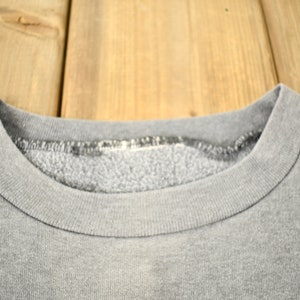 Puede incluir: Primer plano de una sudadera gris con cuello redondo. El cuello y el escote son visibles, mostrando la textura y las costuras de la tela. La sudadera parece vintage o usada, con un aspecto ligeramente descolorido.
