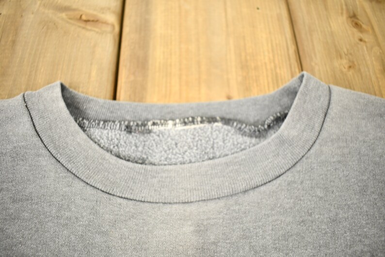 Puede incluir: Primer plano de una sudadera gris con cuello redondo. El cuello y el escote son visibles, mostrando la textura y las costuras de la tela. La sudadera parece vintage o usada, con un aspecto ligeramente descolorido.
