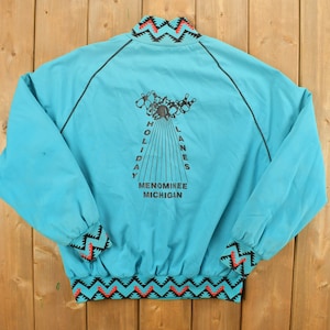 May include: A turquoise blue jacket with black piping and a black, red, and turquoise patterned trim. The back of the jacket features a graphic of bowling pins and a ball with the text "Holiday Lanes Menominee Michigan".