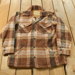 May include: A vintage brown plaid jacket with two chest pockets and two lower pockets. The jacket has a button-down front and buttoned cuffs. The collar has a small tag. The jacket is made of a thick, textured fabric.