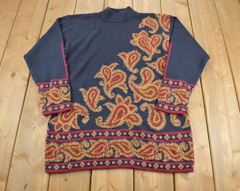 Puede incluir: Un suéter azul marino con un patrón de paisley en tonos de marrón, rojo y dorado. El suéter tiene cuello alto y mangas largas.
