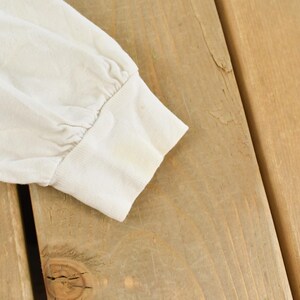 May include: Close-up of a cream-colored long-sleeved shirt sleeve. The cuff is visible, showing slight discoloration. The sleeve is made of a soft, possibly cotton, material. The background is a light brown wooden surface.