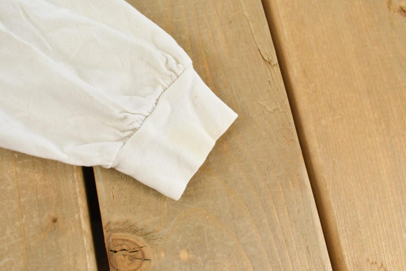 May include: Close-up of a cream-colored long-sleeved shirt sleeve. The cuff is visible, showing slight discoloration. The sleeve is made of a soft, possibly cotton, material. The background is a light brown wooden surface.