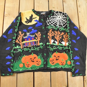 Könnte beinhalten: Schwarze Halloween-Cardigan-Strickjacke mit Knopfverschluss. Mit gestickten orangefarbenen Kürbissen, einem Spukhaus, einer Hexe auf einem Besen und einem weißen Spinnennetz-Design. Grüne Ranken und blaue Fledermausformen verzieren den Pullover.