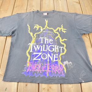 May include: Gray t-shirt featuring the text "The Twilight Zone Tower of Terror" in white, purple, and yellow. The design includes a stylized image of a tower with lightning bolts. The shirt is made of a soft material.