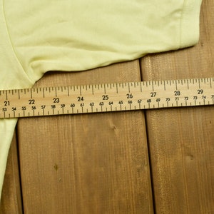 K&ouml;nnte beinhalten: Nahaufnahme eines gelben T-Shirts, das auf einer Holzoberfl&auml;che liegt. Ein Lineal liegt auf dem Shirt und zeigt die Ma&szlig;e in Zoll.