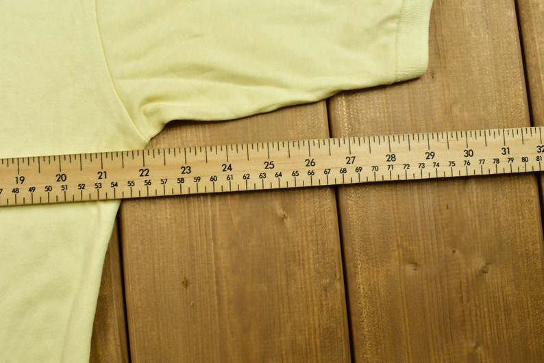 K&ouml;nnte beinhalten: Nahaufnahme eines gelben T-Shirts, das auf einer Holzoberfl&auml;che liegt. Ein Lineal liegt auf dem Shirt und zeigt die Ma&szlig;e in Zoll.