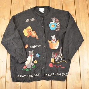 May include: Black button-up cardigan sweater with embroidered cat designs. The sweater features various cat illustrations, the word "MEOW", and the phrase "A CAT IS A CAT".