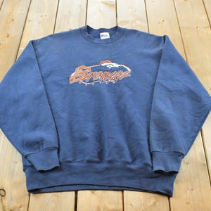May include: A navy blue crewneck sweatshirt featuring the Denver Broncos logo embroidered in orange and white on the chest. The long-sleeve pullover has ribbed cuffs and a ribbed hem, with a visible brand tag located at the inner neckline.
