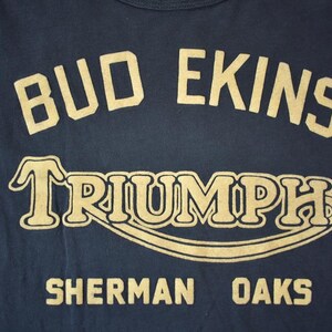 May include: A navy blue t-shirt with gold lettering that reads "Bud Ekins Triumph Sherman Oaks".
