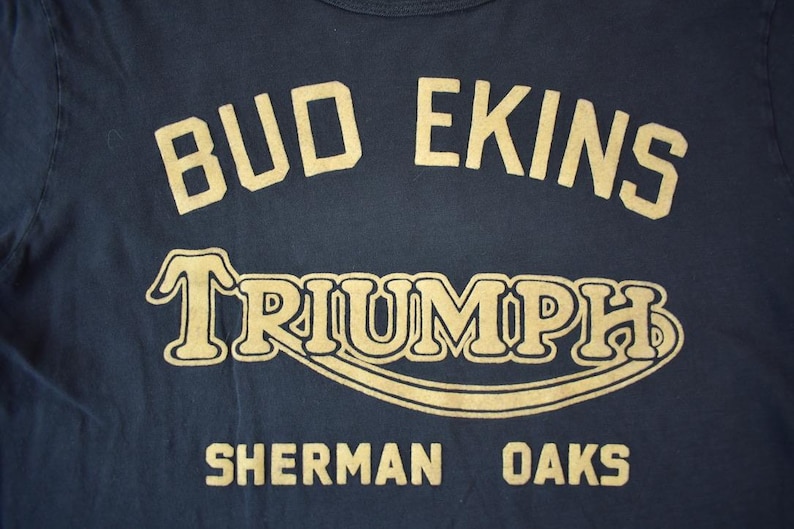 May include: A navy blue t-shirt with gold lettering that reads "Bud Ekins Triumph Sherman Oaks".