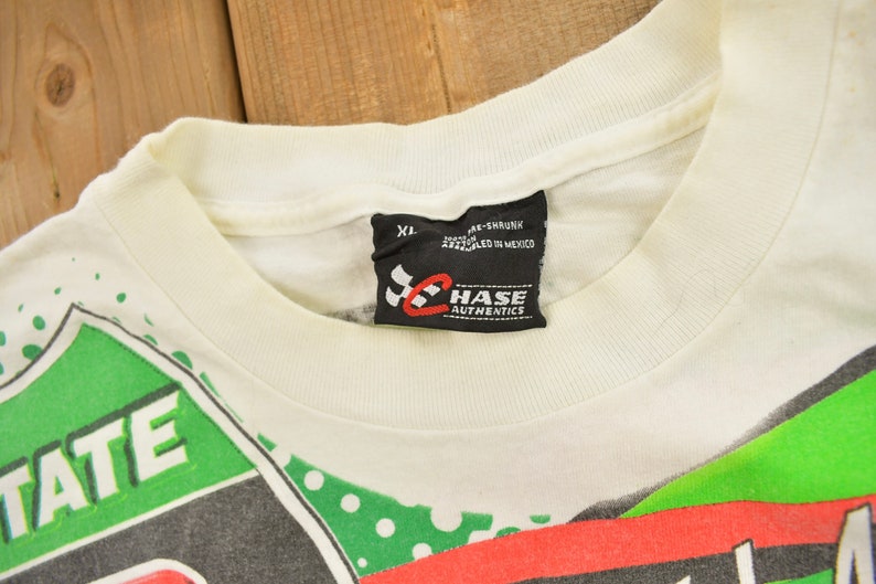 May include: White t-shirt with a green, red and black graphic print featuring the text "STATE" and a chequered flag. The shirt has a black tag with the text "CHASE AUTHENTICS" and the size "XL".