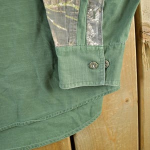May include: Close-up of a green button-down shirt with a camouflage pattern on the upper sleeve. The shirt has two buttons on the cuff and a curved hem. The fabric has a subtle vertical texture.