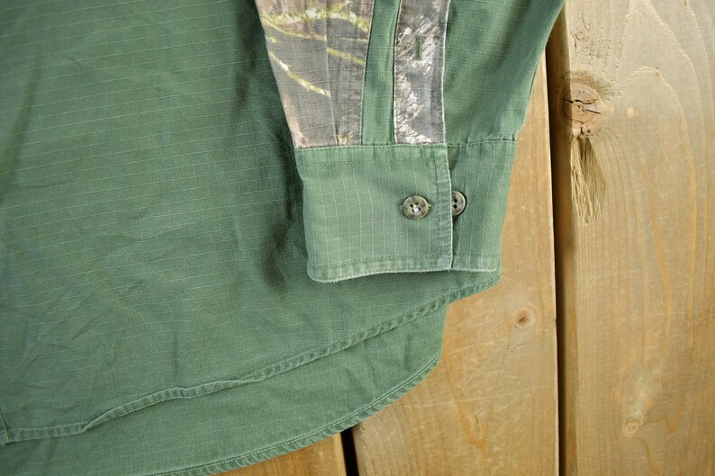 May include: Close-up of a green button-down shirt with a camouflage pattern on the upper sleeve. The shirt has two buttons on the cuff and a curved hem. The fabric has a subtle vertical texture.