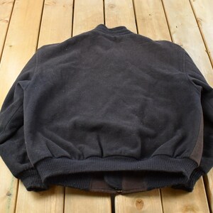 May include: A solid black bomber jacket featuring a ribbed collar, ribbed cuffs, and a ribbed hem. The jacket is constructed from a textured fabric and includes a dark brown panel detail along the side, showcasing a simple and functional design.