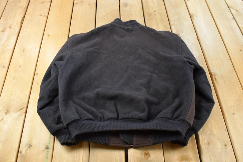 May include: A solid black bomber jacket featuring a ribbed collar, ribbed cuffs, and a ribbed hem. The jacket is constructed from a textured fabric and includes a dark brown panel detail along the side, showcasing a simple and functional design.