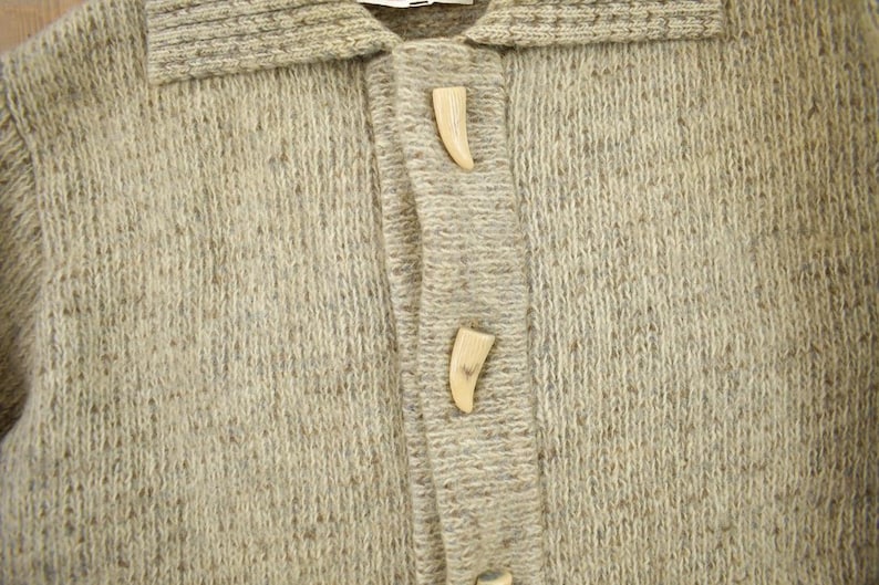 May include: Close-up of a light brown cardigan sweater with a collar and two decorative horn-shaped buttons. The sweater is made of a textured knit fabric.