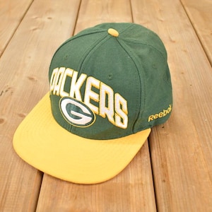 May include: Green and yellow baseball cap with the word "PACKERS" in white block letters. The cap has a yellow brim and a green crown. The Reebok logo is visible on the side.