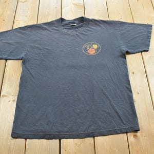 Pode incluir: Uma t-shirt cinzento-carvão de manga curta com um pequeno gráfico circular na parte superior esquerda do peito. O gráfico contém uma ilustração amarela e laranja de um sol, uma lua e estrelas, com texto pequeno a rodear o design.