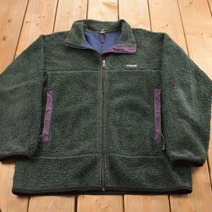 May include: A dark green fleece jacket with a full zipper, featuring purple accents on the pockets and collar. The jacket has a blue inner lining and a small logo on the chest. The jacket is laid flat on a wooden surface.