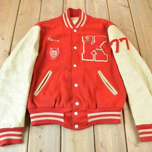 Puede incluir: Chaqueta de béisbol roja y blanca con una manga de cuero blanco, un cuerpo de lana roja y una gran letra blanca "K" en la parte delantera. La chaqueta tiene un amuleto de diablo rojo en la manga izquierda y el número "77" en la manga derecha. El nombre "Dave" está bordado en la manga izquierda.
