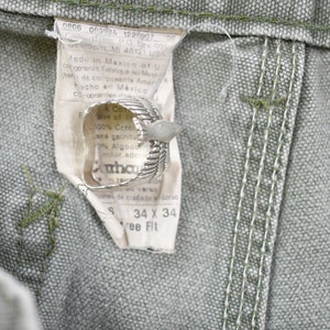 May include: A close-up of a green workwear fabric with a white tag attached. The tag has text that reads "Made in Mexico of US components Fabriqu&eacute; au Mexique de composants Am&eacute;ricains Hecho en M&eacute;xico Componentes". The tag also has a size label that reads "S 34 X 34 Free Fit".