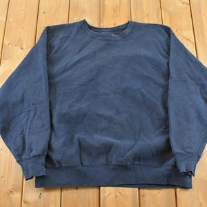 Puede incluir: Sudadera azul marino de cuello redondo y manga larga. La sudadera tiene dobladillo y puños acanalados. La prenda está colocada sobre una superficie de madera.