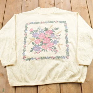 Puede incluir: Una sudadera de manga larga de color crema con un estampado floral dentro de un marco verde y azul. Las flores son rosas, moradas y blancas.
