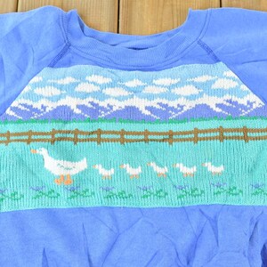 Puede incluir: Una sudadera azul con un dise&ntilde;o tejido de un campo verde, una valla marr&oacute;n, nubes blancas y gansos blancos.