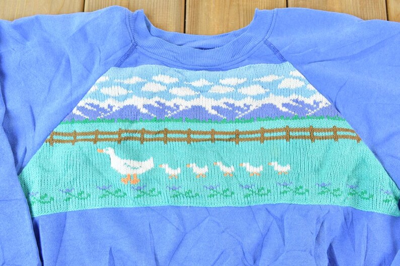 Puede incluir: Una sudadera azul con un dise&ntilde;o tejido de un campo verde, una valla marr&oacute;n, nubes blancas y gansos blancos.