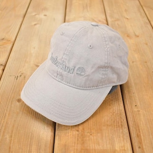 Op de afbeelding: Lichtgrijze baseballcap met het geborduurde woord "Timberland" op de voorkant. De cap heeft een gebogen klep en een klein boomlogo. De hoed is gemaakt van een zacht materiaal.