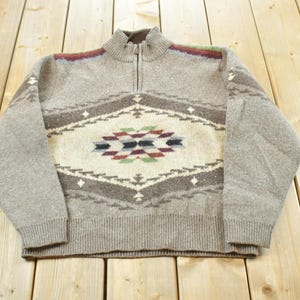 Peut inclure: Un pull zipp&eacute; gris clair avec un motif g&eacute;om&eacute;trique dans les tons marron, cr&egrave;me et des touches de rouge, vert et bleu. Le pull a un col et des poignets c&ocirc;tel&eacute;s. Le motif est centr&eacute; sur la poitrine.