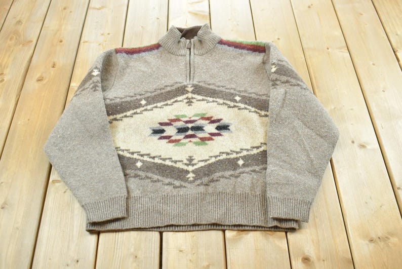 Peut inclure: Un pull zipp&eacute; gris clair avec un motif g&eacute;om&eacute;trique dans les tons marron, cr&egrave;me et des touches de rouge, vert et bleu. Le pull a un col et des poignets c&ocirc;tel&eacute;s. Le motif est centr&eacute; sur la poitrine.