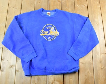 Könnte beinhalten: Ein blaues Sweatshirt mit einer gelben und weißen Grafik, auf der "New York" in einem Kreis steht.
