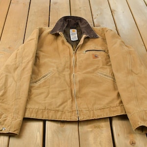 Peut inclure: Une veste de travail en toile beige avec un col en velours côtelé marron foncé, une fermeture éclair intégrale et une petite poche poitrine zippée. La veste présente une étiquette Carhartt sur la poitrine, deux poches avant et des poignets boutonnés.