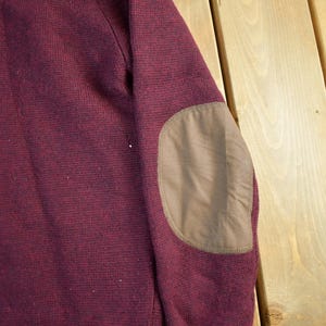 Puede incluir: Primer plano de una manga de su&eacute;ter burdeos con un parche en el codo marr&oacute;n. El su&eacute;ter tiene un patr&oacute;n de punto texturizado. El parche del codo es de un material liso y liso. El fondo es una superficie de madera de color claro.