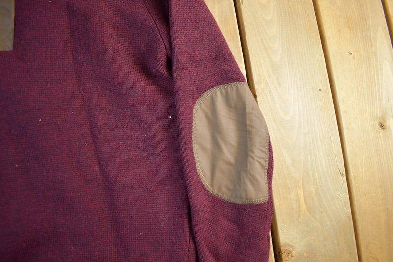 Puede incluir: Primer plano de una manga de su&eacute;ter burdeos con un parche en el codo marr&oacute;n. El su&eacute;ter tiene un patr&oacute;n de punto texturizado. El parche del codo es de un material liso y liso. El fondo es una superficie de madera de color claro.