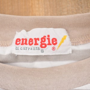 Może przedstawiać: Biało-beżowa w paski koszulka z białą metką z napisem "energie by currants" czerwonymi literami, ż&oacute;łtym błyskawicą i symbolem znaku towarowego.