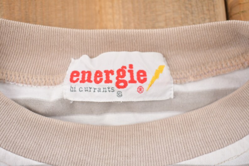 Może przedstawiać: Biało-beżowa w paski koszulka z białą metką z napisem "energie by currants" czerwonymi literami, ż&oacute;łtym błyskawicą i symbolem znaku towarowego.
