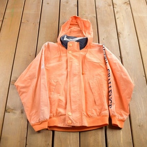 Puede incluir: Una chaqueta con capucha de color melocotón con una cremallera azul marino y una impresión "URBAN CHALLENGE" azul marino en la manga derecha.