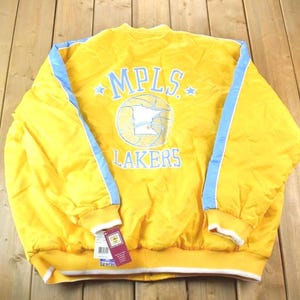 Pode incluir: Casaco amarelo vintage com detalhes em azul e o texto "MPLS LAKERS" nas costas. O casaco tem gola, punhos e cós canelados. Uma etiqueta está presa na parte inferior. O casaco é feito de um material brilhante.