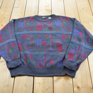 Peut inclure: Un pull à manches longues et col rond avec une base gris anthracite et un motif géométrique multicolore dans des tons de rouge, bleu et vert. Le vêtement en tricot présente des poignets et un ourlet côtelés, avec un motif à rayures horizontales.