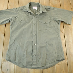 Peut inclure: Chemise vert olive &agrave; manches courtes avec deux poches de poitrine et une fermeture boutonn&eacute;e. La chemise a un col et des &eacute;paulettes sur les &eacute;paules. Le tissu semble &ecirc;tre l&eacute;ger.